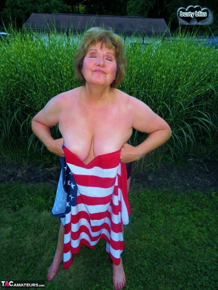 Older plumper Busty Bliss holds an American flag while totally naked in a yard - #1