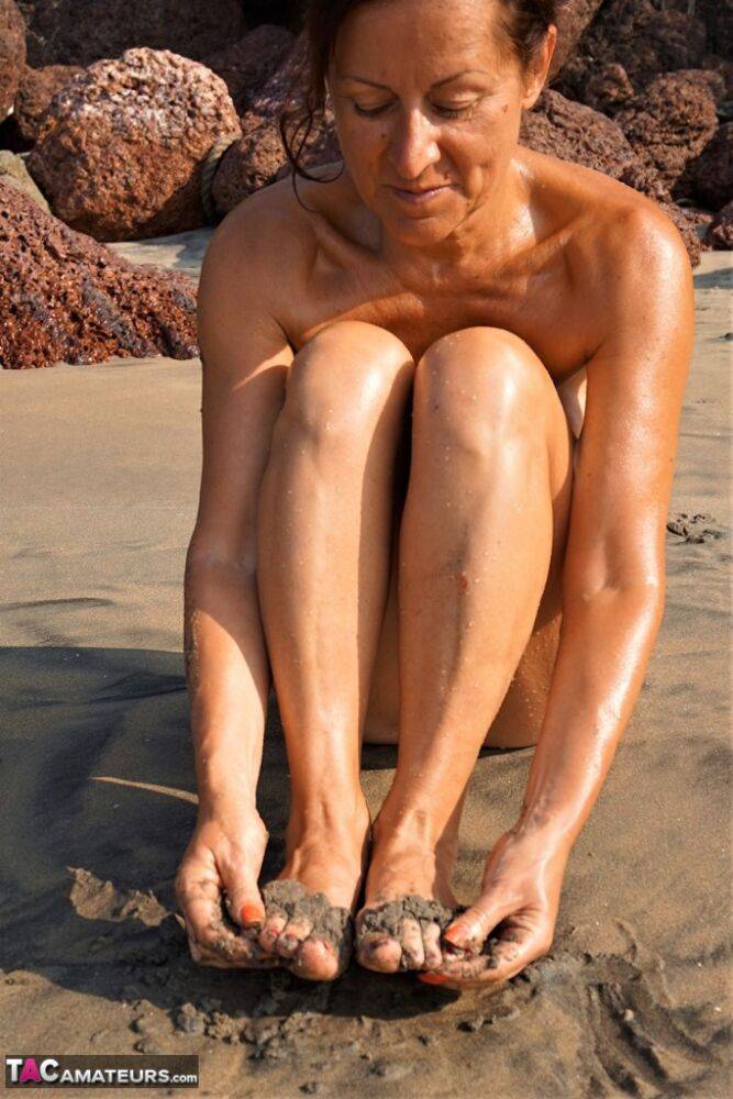 Naked older woman Diana Ananta covers her feet in beach sand at low tide - #9