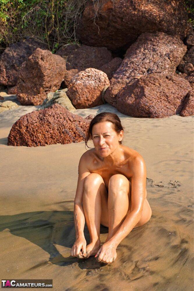 Naked older woman Diana Ananta covers her feet in beach sand at low tide - #11