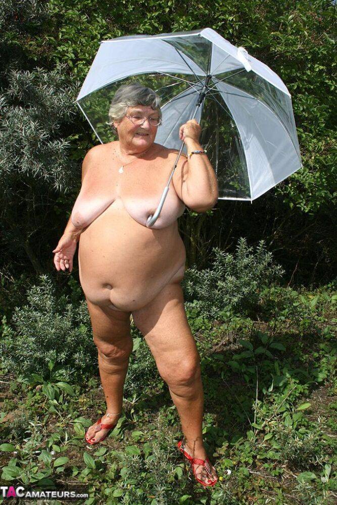 Obese oma Grandma Libby holds an umbrella while posing naked by fir trees - #10